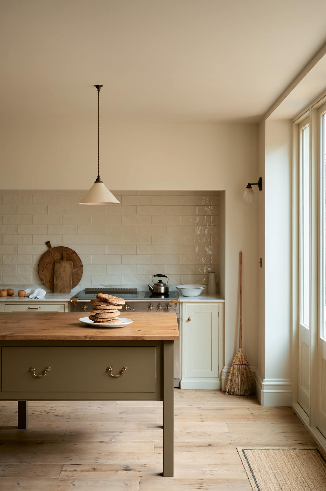A light, clean, and simple shaker kitchen features a sink and cooker run nestled in a chimney breast, cream porcelain splashback tiles, wooden floorboards, and white walls. Cream cabinets complement an olive green Georgian-style prep table with deep drawers and a rustic oak worktop at the room’s centre, with an elegant creamware pendant light.