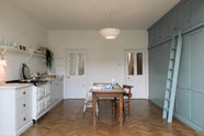A beautiful Trinity Blue Shaker kitchen featuring a cooker and sink run, floor-to-ceiling pantry cabinets with a matching ladder, and a vintage wooden dining table with a wonderfully worn top. A long Shaker shelf spans above the kitchen area, complemented by a white scalloped porcelain chandelier and grand solid oak herringbone floors.