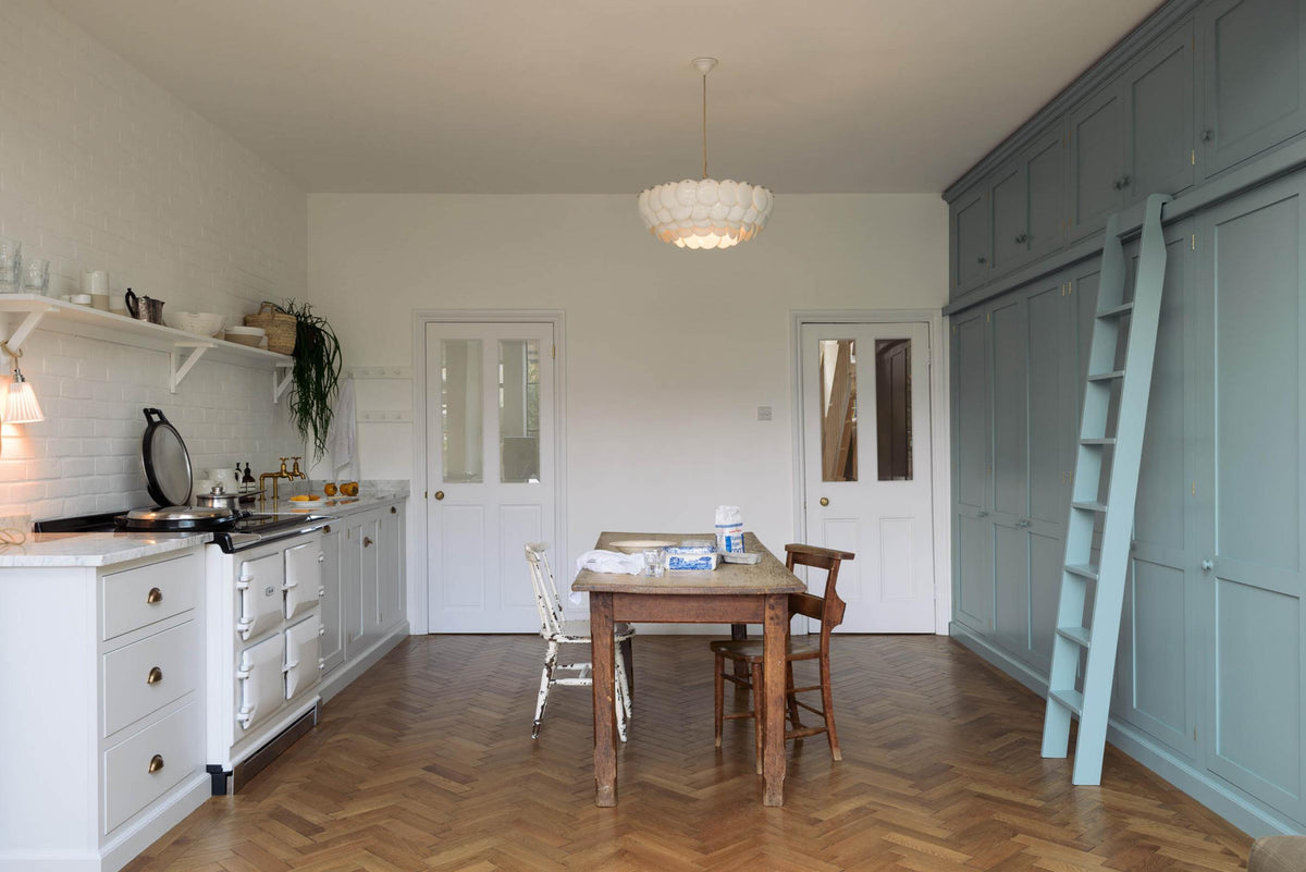 A beautiful Trinity Blue Shaker kitchen featuring a cooker and sink run, floor-to-ceiling pantry cabinets with a matching ladder, and a vintage wooden dining table with a wonderfully worn top. A long Shaker shelf spans above the kitchen area, complemented by a white scalloped porcelain chandelier and grand solid oak herringbone floors.