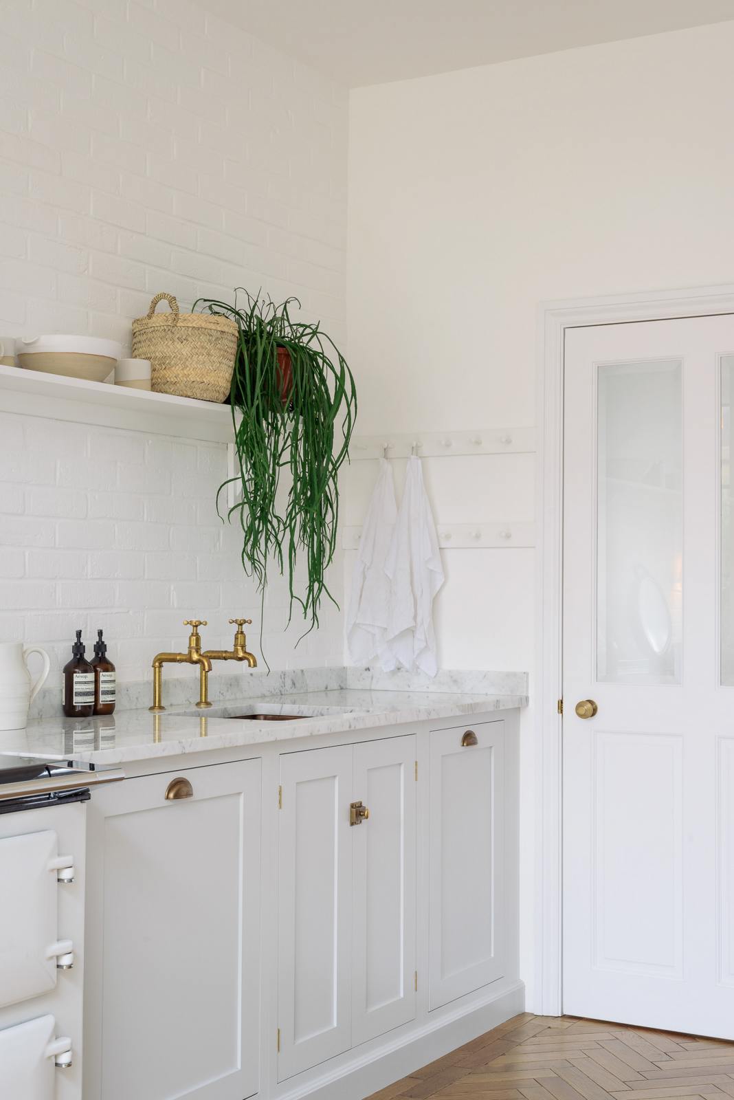 An oblique view of a beautiful Trinity Blue Shaker kitchen with a cooker and sink run, a long Shaker shelf overhead, peg rails, a white scalloped porcelain chandelier, and grand solid oak herringbone floors.