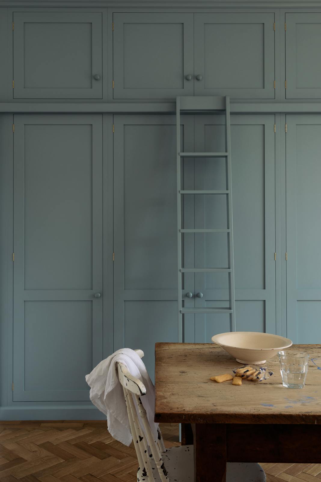 A close-up of a beautiful Trinity Blue Shaker kitchen featuring floor-to-ceiling pantry cabinets with a matching ladder, and a vintage wooden dining table with a wonderfully worn top. The table has a collection of baking equipment and ingredients.
