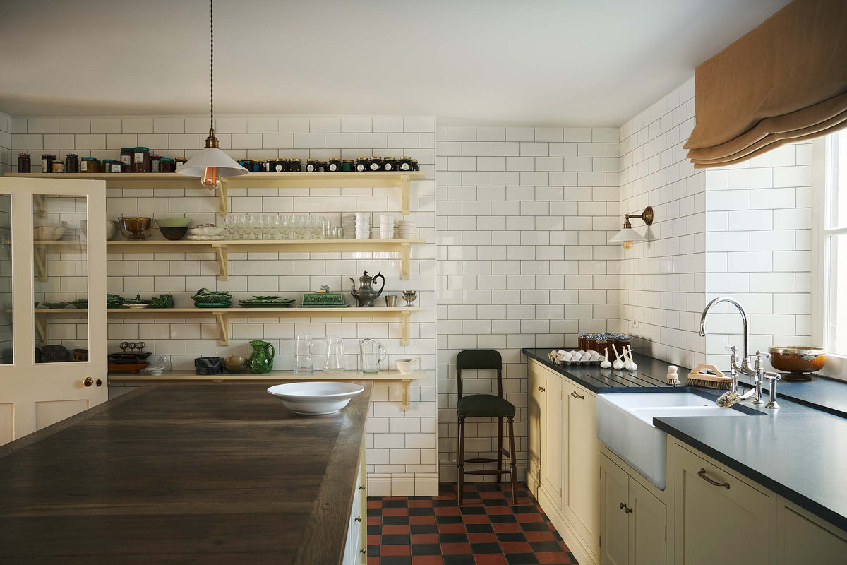A landscape shot of a moody and atmospheric butter-yellow painted shaker kitchen with a simple, utilitarian feel. Light cupboards and glossy, white tiled walls contrast with original black and red chequerboard quarry tiles. The scullery features a central island, a long run of base cabinets that includes a double sink unit, honed back granite worktops and fitted Shaker shelving.