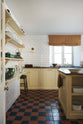 A simple run of Shaker base cupboards and central island, all painted in a buttery yellow hue are topped with honed black granite and reclaimed timber. Glossy white subway tiles cover the walls floor to ceiling, while the floor features original red and black quarry tiles. To the left, four rows of Shaker pantry shelves hold jars of jam, glassware, bowls, and dark green majolica leaf pottery.