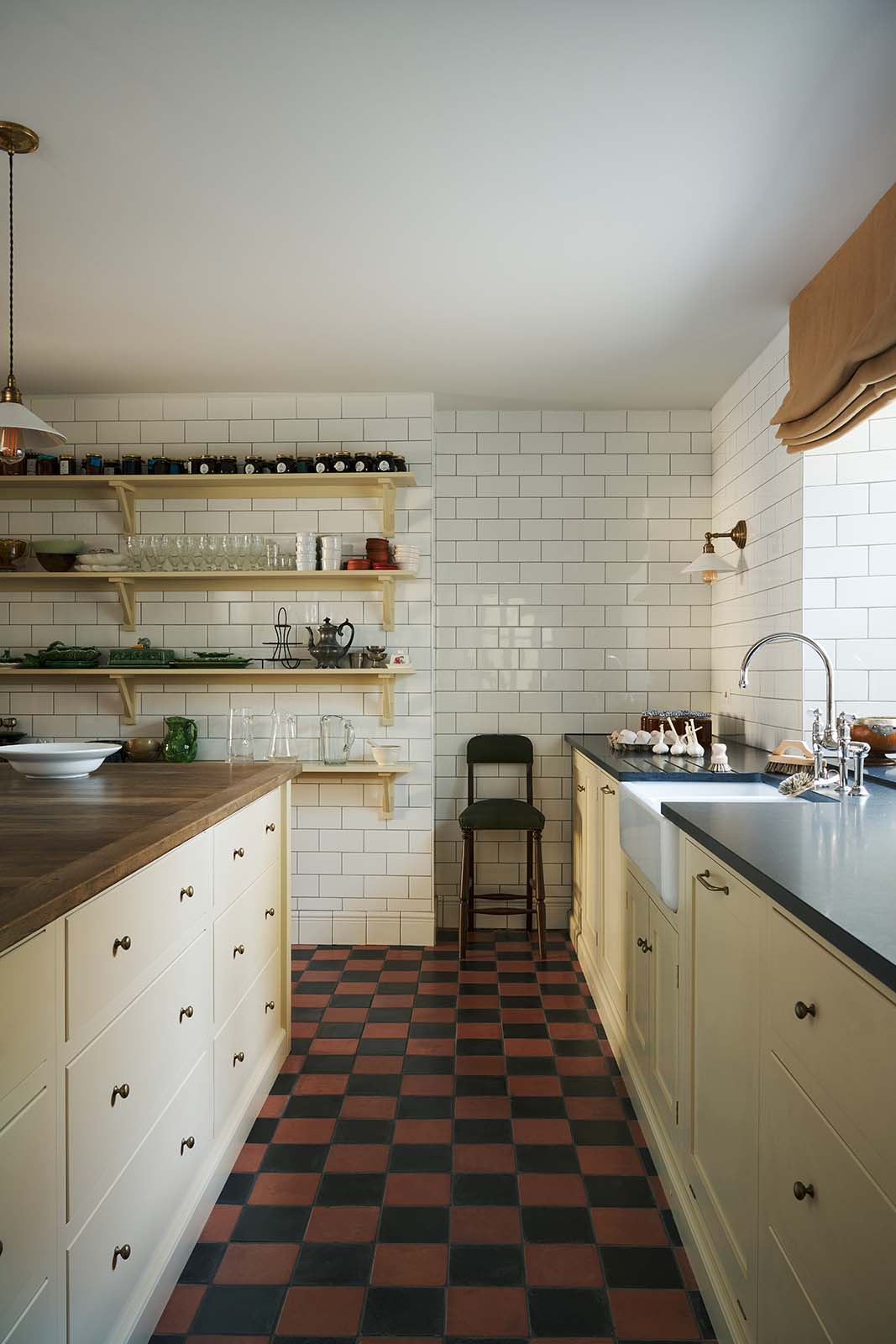 A moody and atmospheric butter-yellow painted shaker kitchen with a simple, utilitarian feel. Light cupboards and glossy, white tiled walls contrast with original black and red chequerboard quarry tiles. The scullery features a central island, a long run of base cabinets that includes a double sink unit, honed back granite worktops and fitted Shaker shelving.