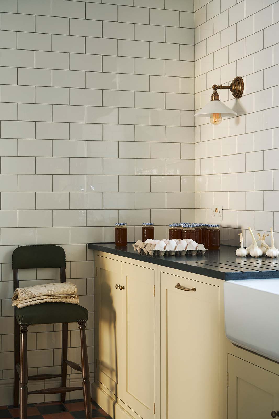 A vignette from the corner of a traditional-style scullery, with pale yellow painted Shaker base cabinets fitted with deVOL’s aged brass knobs and handles. Jars of jam, trays of eggs and fresh garlic bulbs are placed on top of the honed black granite worktop, and an antique green leather barstool is propped up.