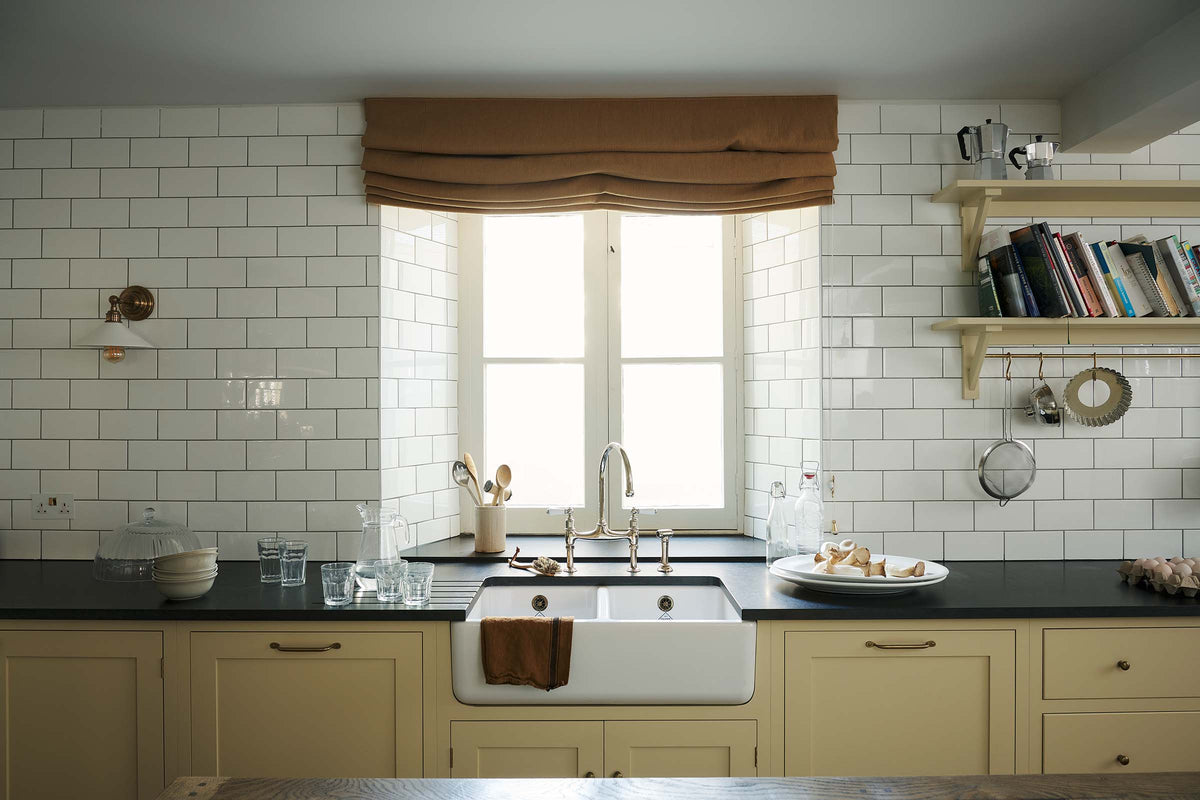 A straight-on view of the sink area in a traditional-style scullery. Pale yellow Shaker base cabinets with white ceramic Belfast sink, honed black granite worktops and glossy white subway tiles create a simple and utilitarian feel.
