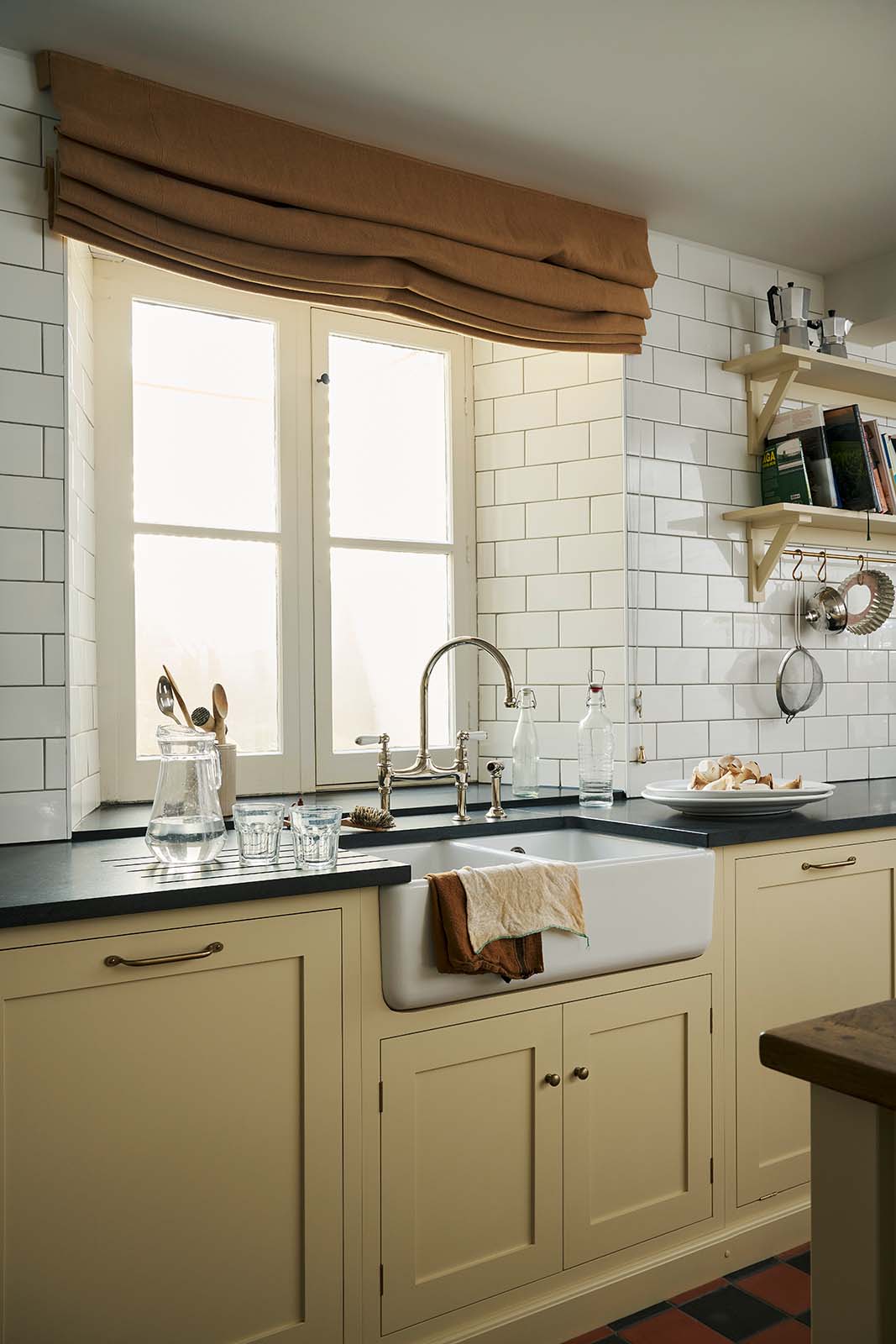 A close-up, oblique shot of the sink area in a traditional-style scullery. Pale yellow Shaker base cabinets with white ceramic Belfast sink, honed black granite worktops and glossy white subway tiles create a simple and utilitarian feel.
