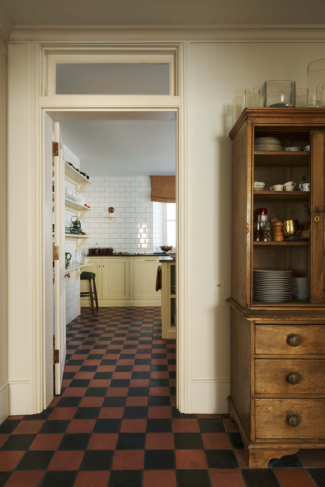 Looking through a doorway into a basement scullery with Shaker base cupboards and a central island, all painted in a soft yellow hue and fitted with a combination of honed black granite and reclaimed wooden worktops. Floor-to-ceiling glossy subway tiles create beautiful reflections, contrasting with the time-worn, original black and red chequered quarry tiles.