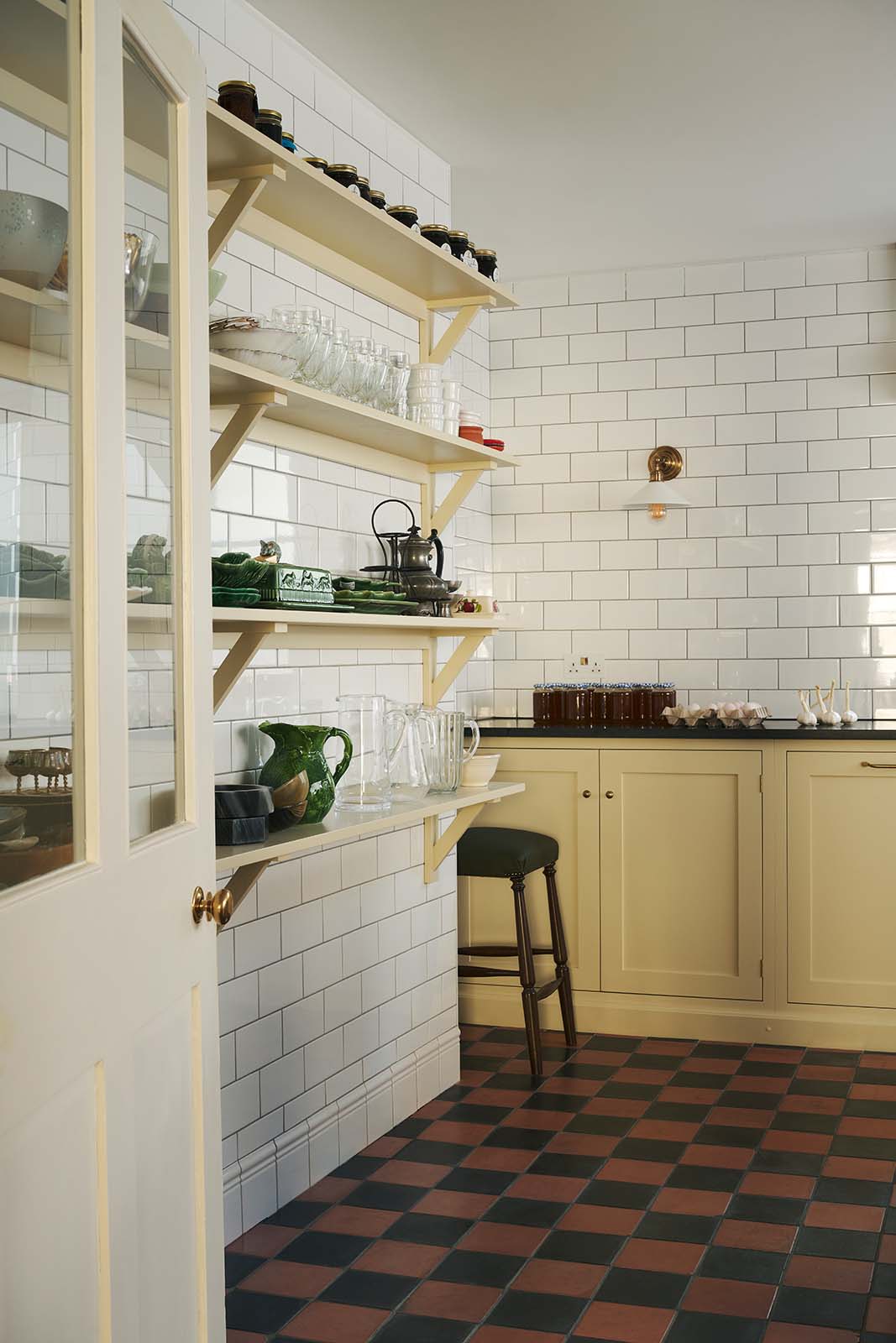 An angled view towards a corner of shaker base cabinets and four fitted shaker shelves, all painted in a soft creamy yellow colour. Glossy white tiled walls and original, black and red quarry tiled floors create a utilitarian feel.