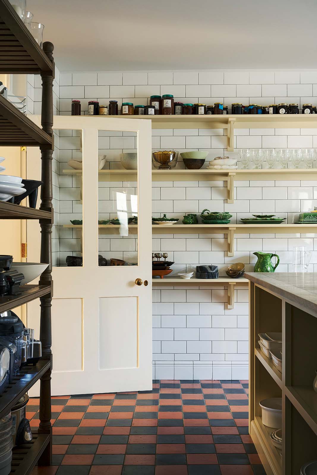 Four rows of Shaker shelves, painted in a muted, buttery yellow hue, are fitted to a white floor-to-ceiling tiled wall, and are stocked with jars of jam, glassware, bowls and vibrant green majolica leaf pottery. This scullery kitchen, found below stairs in a grand Surrey country house, feels very utilitarian with glossy subway tiles and original Victorian red and black chequered quarry tiles.