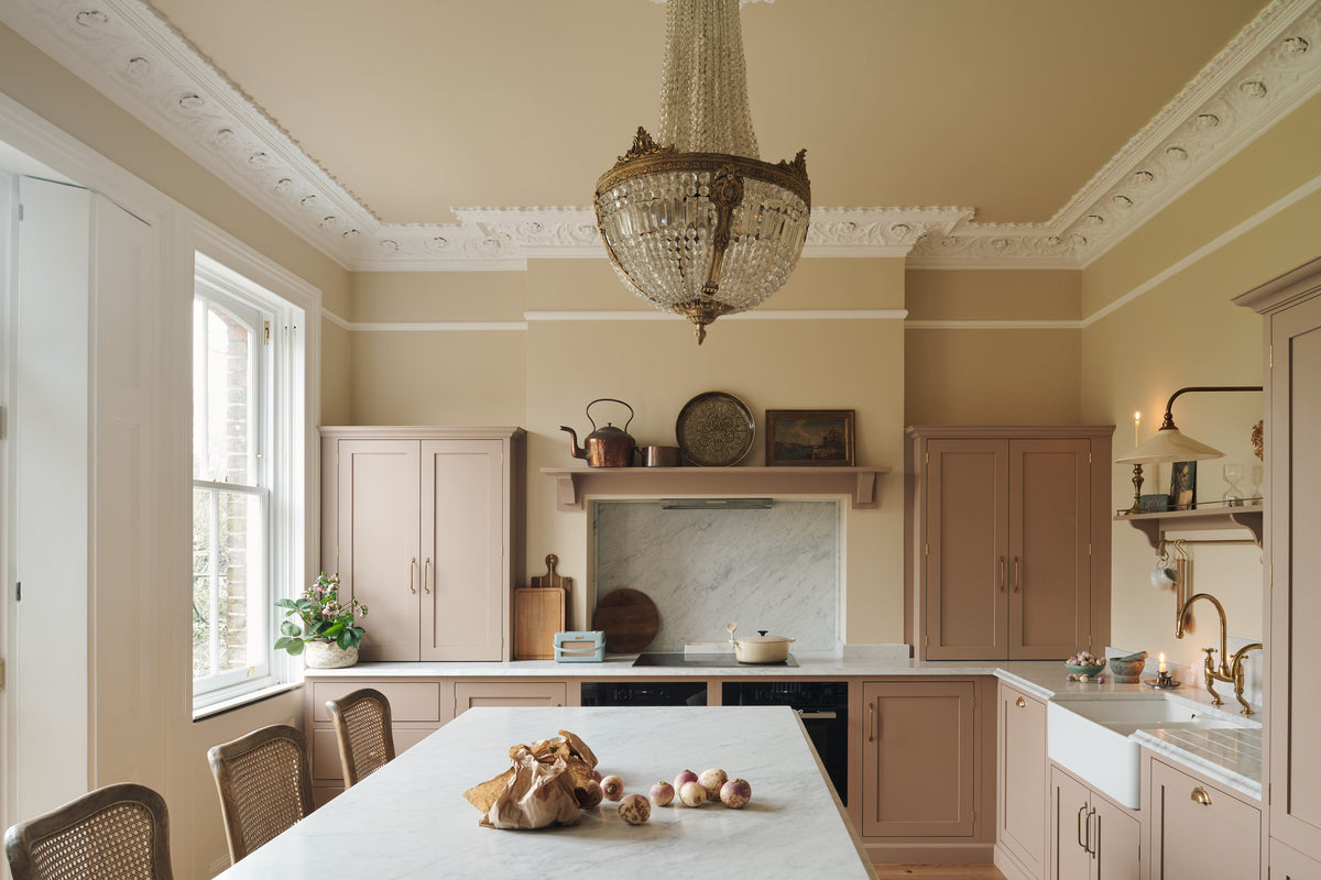 A romantic and peaceful Princelet pink Shaker kitchen features an L-shaped sink and cooker run with a marble worktop, matching countertop cupboards, a kitchen island with a breakfast nook and an overmantle above the chimney breast. Beautifully paired with the subtle butter yellow painted walls, decorative cornice and a grand antique chandelier, it creates a warm and inviting space.