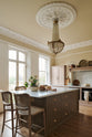 An oblique view of a romantic and peaceful Princelet pink Shaker kitchen features an L-shaped sink and cooker run with a marble worktop, matching countertop cupboards, a kitchen island with a breakfast nook and an overmantle above the chimney breast. Beautifully paired with the subtle butter yellow painted walls, decorative cornice and a grand antique chandelier, it creates a warm and inviting space.