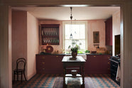 A warm and simply gorgeous refectory-red Shaker kitchen features a sink run with a black granite worktop, a wall cupboard, a small Heirloom plate rack, a pantry cupboard, and a gas stove in a chimney breast with Sepia Lace market splashback tiles. Beautifully decorated with vintage furniture, artisan ceramics, original botanical illustrations, reeded glass pendant lights, and a wild floral arrangement, it creates a humble and endearing space.