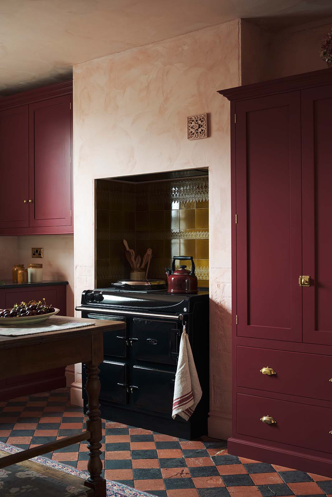 An oblique view of a warm and simply gorgeous refectory-red Shaker kitchen features a sink run with a black granite worktop, a wall cupboard, a pantry cupboard, and a gas stove in a chimney breast with Sepia Lace market splashback tiles. Beautifully decorated with vintage furniture, artisan ceramics, and a matching red teapot, it creates a humble and endearing space.