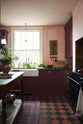 A warm and simply gorgeous refectory-red Shaker kitchen features a sink run with a black granite worktop, a wall cupboard, a small Heirloom plate rack, a wall cupboard, a pantry cupboard, and a gas stove in a chimney breast with Sepia Lace market splashback tiles. Beautifully decorated with vintage furniture, artisan ceramics, original botanical illustrations, reeded glass pendant lights, and a wild floral arrangement, it creates a humble and endearing space.