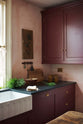 A closer look at a warm and simply gorgeous refectory-red Shaker kitchen features a sink run with a black granite worktop and a wall cupboard. Beautifully decorated with vintage furniture, artisan ceramics, original botanical illustrations, and a healthy bunch of rosemary, it creates a humble and endearing space.
