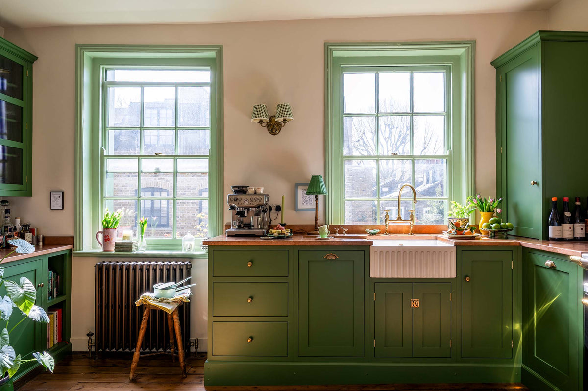 A bright and humble lime-green kitchen with an L-shaped cooker and sink run topped with aged copper worktops, featuring countertop and glazed cupboards, a single fluted Belfast sink with an aged brass Ionian tap, and colourful accents like tulips, a bowl of limes, macaroons, and assorted bottles of alcohol.