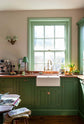 A closer look at a bright and humble lime-green kitchen with an L-shaped cooker and sink run topped with aged copper worktops, a countertop and glazed cupboards, a single fluted Belfast sink with an aged brass Ionian tap, and colourful accents like tulips, a bowl of limes and macaroons. 