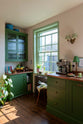 A corner of a bright and humble lime-green kitchen with an L-shaped cooker and sink run topped with aged copper worktops, a glazed counter cabinet, and colourful accents like tulips, a bowl of lemons, macaroons, books and assorted bottles of alcohol.