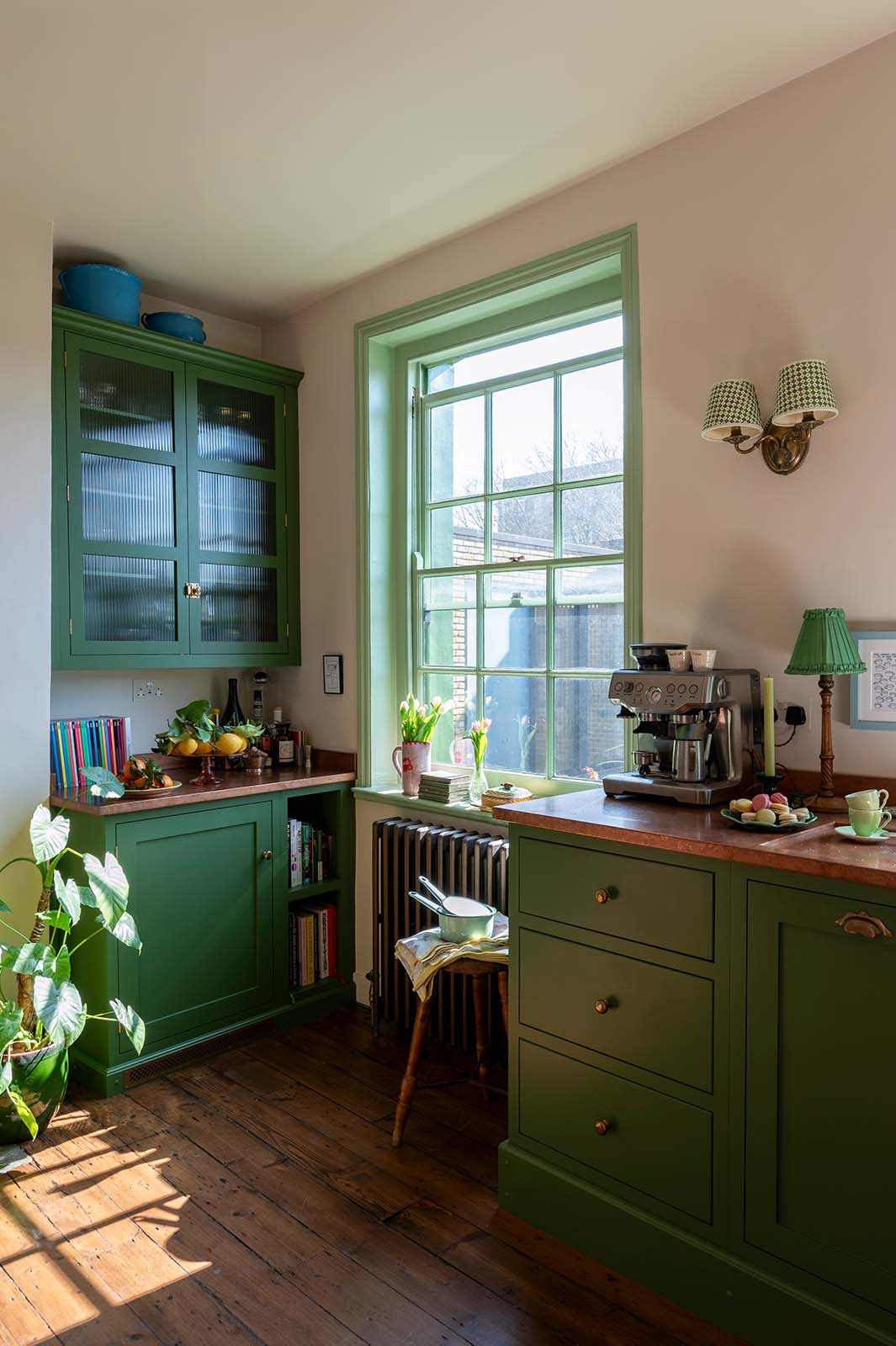 A corner of a bright and humble lime-green kitchen with an L-shaped cooker and sink run topped with aged copper worktops, a glazed counter cabinet, and colourful accents like tulips, a bowl of lemons, macaroons, books and assorted bottles of alcohol.