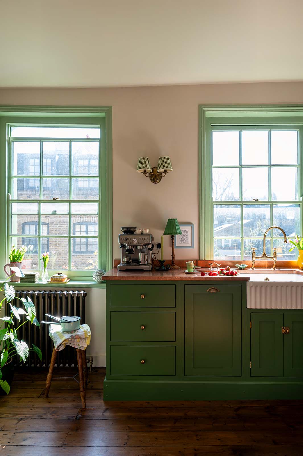 A bright and humble lime-green kitchen with an L-shaped cooker and sink run topped with aged copper worktops, a single fluted Belfast sink with an aged brass Ionian tap, and colourful accents like tulips, a plate of strawberries, mint tea cups and a silver coffee machine.