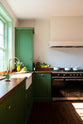 An angled view of a bright and humble lime-green kitchen with an L-shaped cooker and sink run topped with aged copper worktops, featuring countertop and glazed cupboards, a shaker canopy, a single fluted Belfast sink with an aged brass Ionian tap, and colourful accents like tulips, a bowl of limes and lemons, mint tea cups, and assorted bottles of wine.
