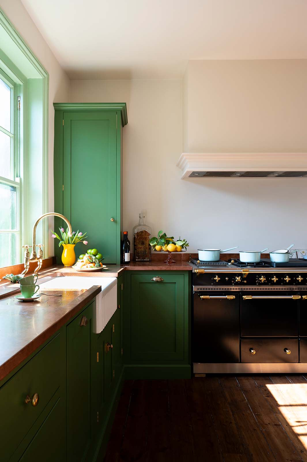 An angled view of a bright and humble lime-green kitchen with an L-shaped cooker and sink run topped with aged copper worktops, featuring countertop and glazed cupboards, a shaker canopy, a single fluted Belfast sink with an aged brass Ionian tap, and colourful accents like tulips, a bowl of limes and lemons, mint tea cups, and assorted bottles of wine.