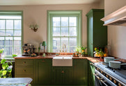 A bright and humble lime-green kitchen with an L-shaped cooker and sink run topped with aged copper worktops, featuring countertop and glazed cupboards, a single fluted Belfast sink with an aged brass Ionian tap, and colourful accents like tulips, a bowl of limes, lemons and oranges, macaroons, and a silver coffee machine.