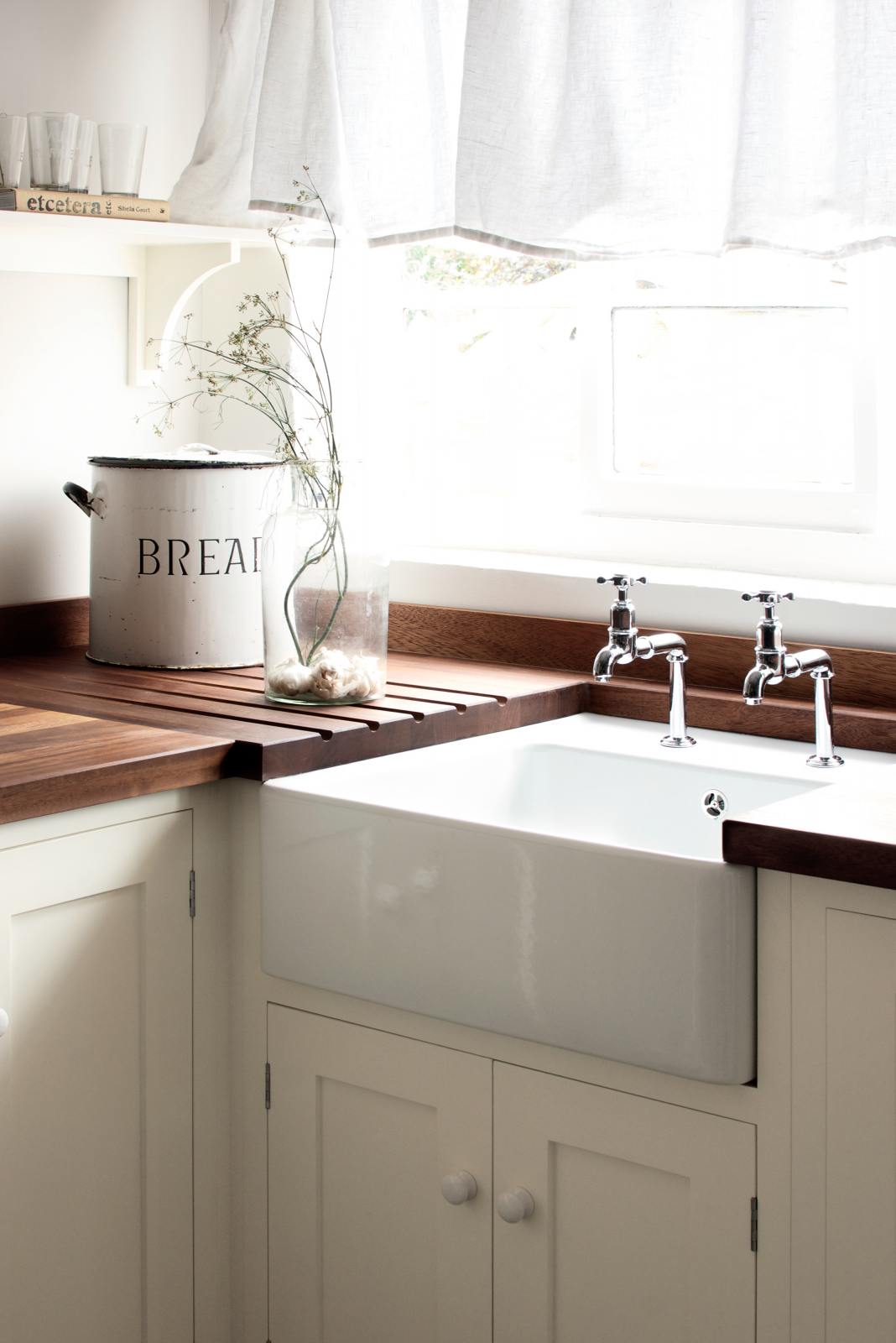 A close look at a sink in a minimalist white linen Shaker kitchen with a dark polished sapele worktop. It is a single sink with antique silver Mayan taps. The kitchen is decorated with a cylinder bread tin, a glass vase with wildflowers and on the shelf is a cook with simple drinking glasses.