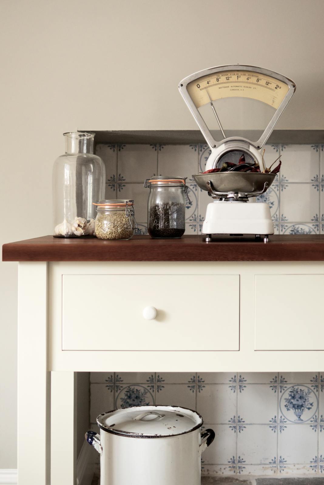 A close-up of a freestanding white linen prep table with a dark polished sapele worktop, styled with glass jars of ingredients, garlic in a glass, and a vintage scale holding chillies. The unused fireplace is finished with Delft-style ceramic splashback tiles.
