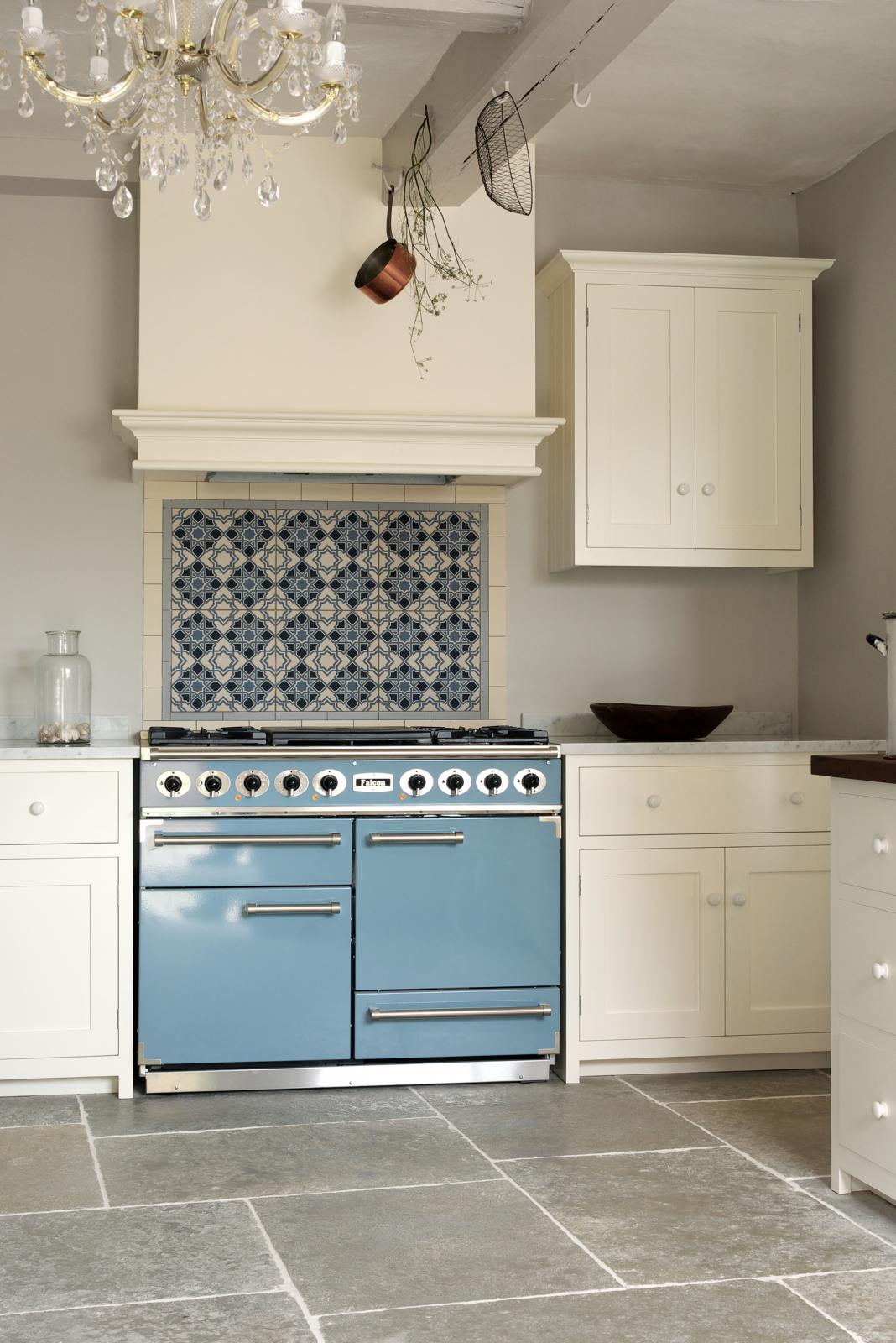 A cooker run in a minimalist white linen Shaker kitchen with a Carrara marble worktop, matching cupboard, and Shaker canopy. A muted blue gas cooker sits beneath a detailed blue floral splashback.
