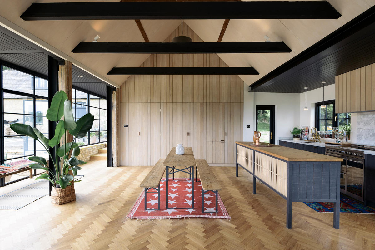 A large and minimal Sebastian Cox kitchen features a long sink and cooker run with a Carrara marble worktop, a single copper sink, an aged brass Ionian tap, a canopy, a built-in bank of cupboards, and a large kitchen island with a solid oak worktop. Beautifully decorated with artisan ceramics, a large indoor plant, a wicker basket, and bright patterned rugs, this kitchen creates a simple and practical space.