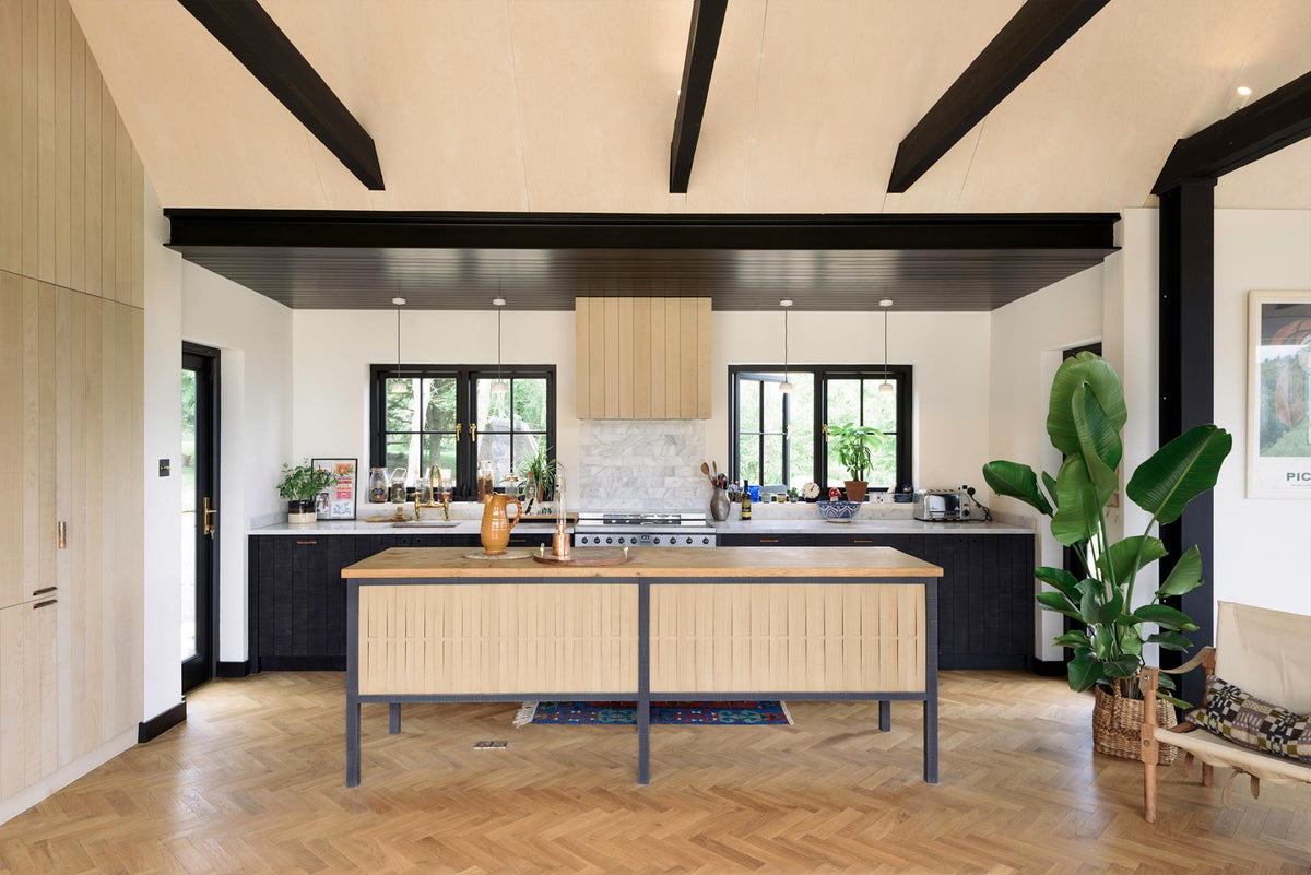 A large and minimal Sebastian Cox kitchen features a long sink and cooker run with a Carrara marble worktop, a single copper sink, an aged brass Ionian tap, a canopy, and a large kitchen island with a solid oak worktop. Beautifully decorated with artisan ceramics, indoor plants, a wicker basket, and bright patterned rugs, this kitchen creates a simple and practical space.