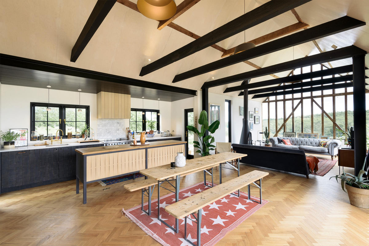 An oblique view of a large and minimal Sebastian Cox kitchen features a long sink and cooker run with a Carrara marble worktop and a large kitchen island with a solid oak worktop. Beautifully decorated with artisan ceramics, a large indoor plant, a vintage park bench dining table, and bright patterned rugs, this kitchen creates a simple and practical space.