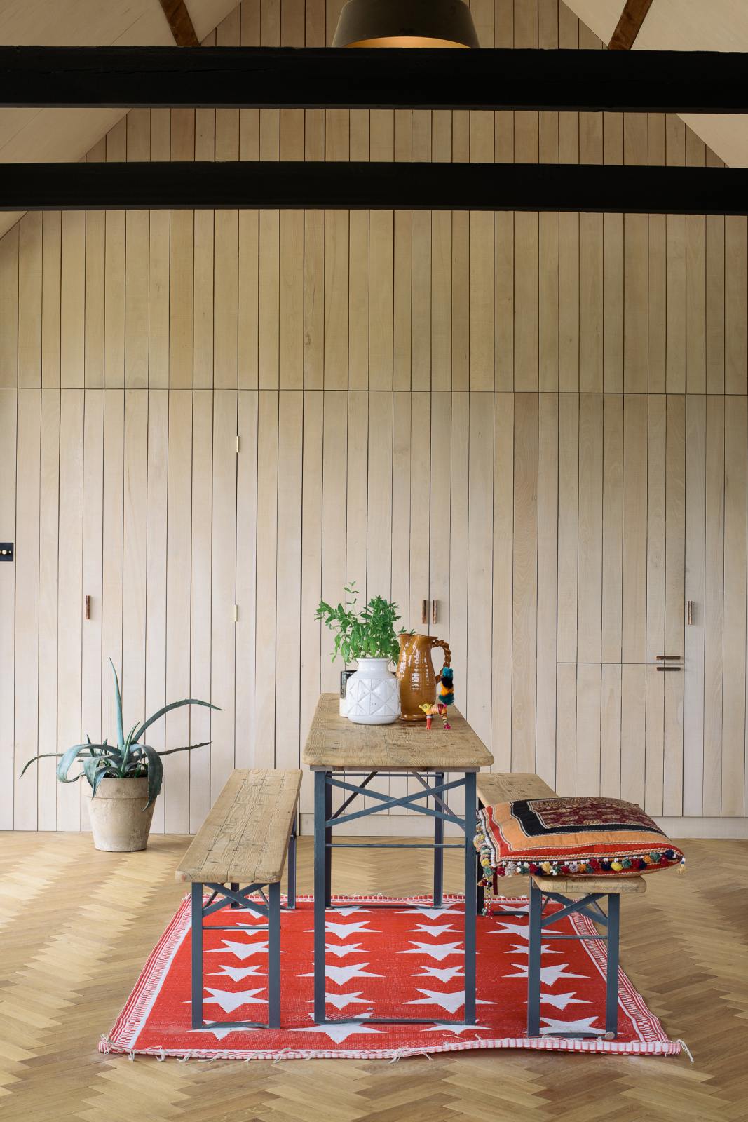 A large and minimal Sebastian Cox kitchen features a built-in bank of cupboards and a vintage park bench dining table. Beautifully decorated with artisan ceramics, a large indoor plant, a quirky little toy, a colourful pillow, and a bright patterned rug, this kitchen creates a simple and practical space.