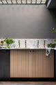 A chic and simple Sebastian Cox basement kitchen features a long sink run with two copper sinks, aged brass Mayan taps, and a Carrara marble worktop and splashback. This kitchen creates a cool and atmospheric space, beautifully decorated with luscious green house plants, vintage items, drinking glasses, a bowl of limes, salt, and parsley.