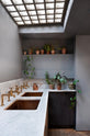 An oblique view of a chic and simple Sebastian Cox basement kitchen features a long sink run with two copper sinks, aged brass Mayan taps, and a Carrara marble worktop and splashback. This kitchen creates a cool and atmospheric space, beautifully decorated with luscious green house plants, a stylish copper watering can, and beautiful ceramic and terracotta pots.