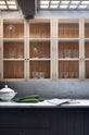 A closer look at a chic and simple Sebastian Cox basement features a long sink run with Carrara marble countertop and splashback, and glazed wall cupboards. This kitchen creates a cool and atmospheric space, beautifully decorated with artisan ceramics, drinking glasses, a book, and bitter melons.