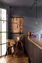 A chic and simple Sebastian Cox basement features a large kitchen island with a gas cooker and a copper and solid oak worktop. This kitchen creates a cool and atmospheric space, beautifully decorated with luscious green cacti, beloved furniture, a Helen stool, a copper pot, a vintage illustration wall-hanging, a carafe of water, a drinking glass, and simple pendant lights.