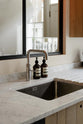 A close-up of a sleek and modern Sebastian Cox galley kitchen’s sink run features a transom window, a stainless steel sink, a silver Quooker tap, hand-wash bottles, and a Carrara marble worktop.