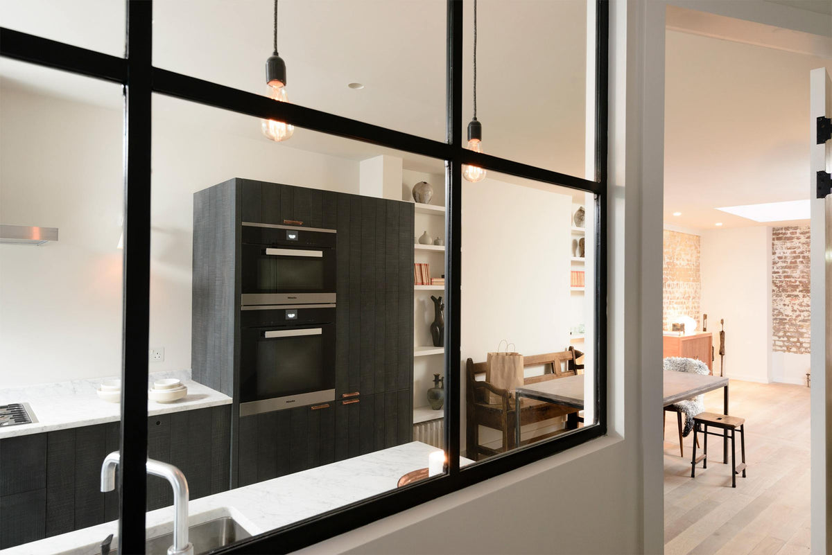 Past a black-framed transom partition window is a sleek and modern Sebastian Cox galley kitchen featuring a sink and cooker run with a Carrara marble worktop, an appliance cabinet, a fridge/freezer cupboard, and a dining space. Adorned with simple ceramics, decorative ceramics, books, bare pendant lights, mismatched vintage dining furniture, and a fluffy wool blanket, this kitchen creates an elegant and practical space.