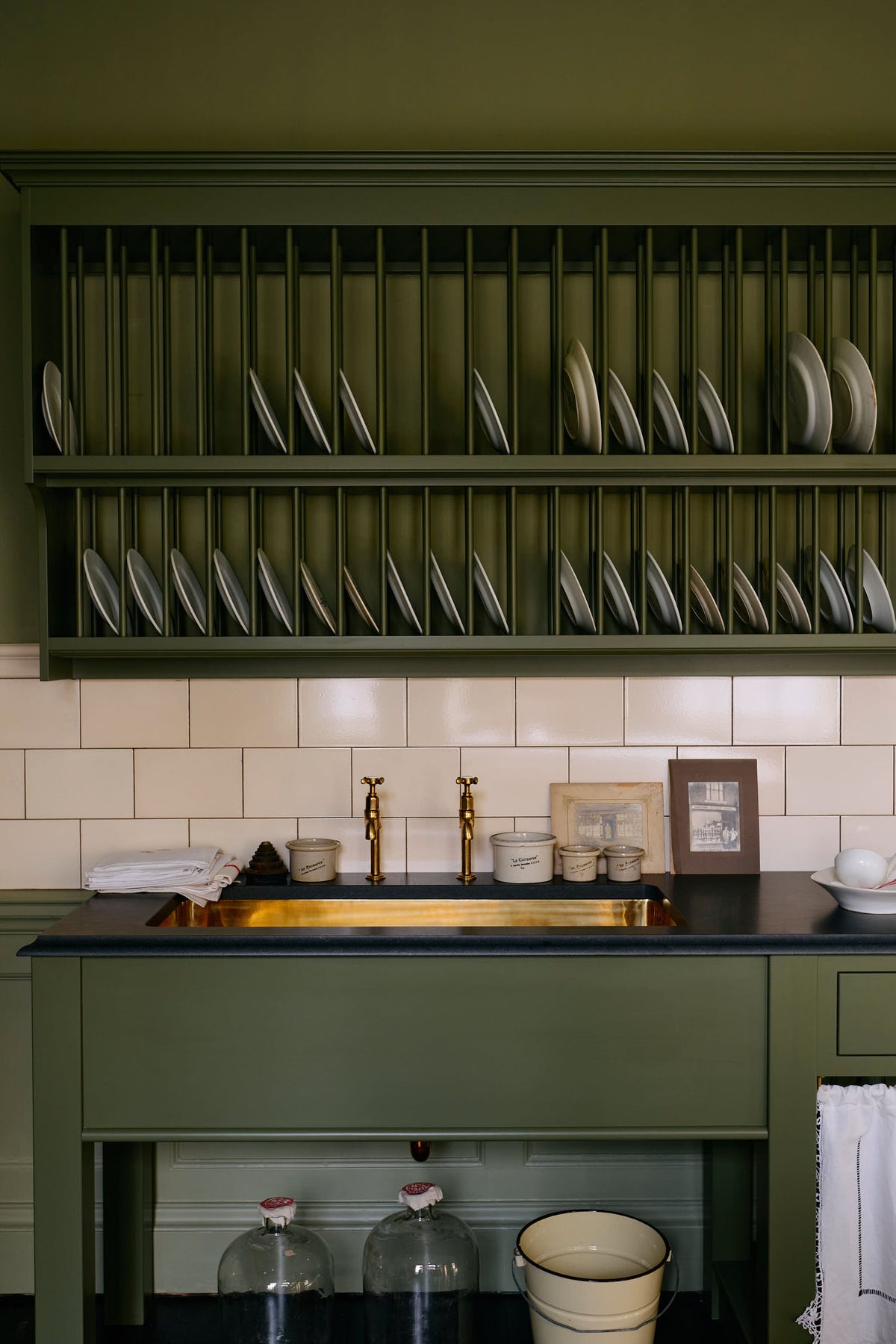 A quaint and stylish Wilkes green Heirloom kitchen features a sink cabinet with a café rail, white lace curtains, a polished brass sink, aged brass Mayan taps, and a large plate rack. Adorned with vintage photos, a cream bucket, simple ceramics, cream subway splashback tiles, and a golden-framed sliced green pumpkin, this kitchen creates a luxurious dining and social space.