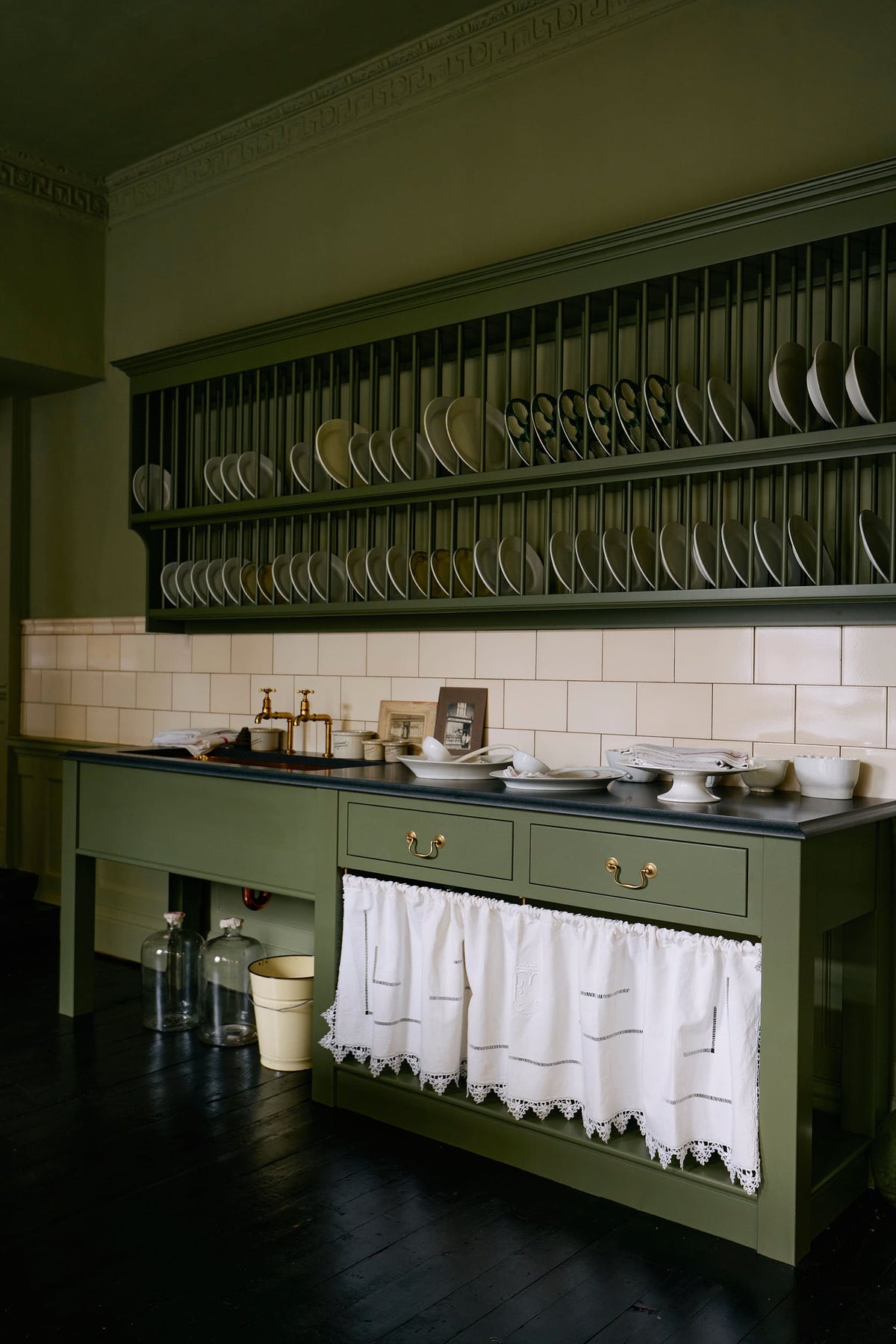 An oblique view of a quaint and stylish Wilkes green Heirloom kitchen features a sink cabinet with a café rail, white lace curtains, a polished brass sink, aged brass Mayan taps, and a large plate rack. Adorned with vintage photos, a cream bucket, simple ceramics, cream subway splashback tiles, and a golden-framed sliced green pumpkin, this kitchen creates a luxurious dining and social space.