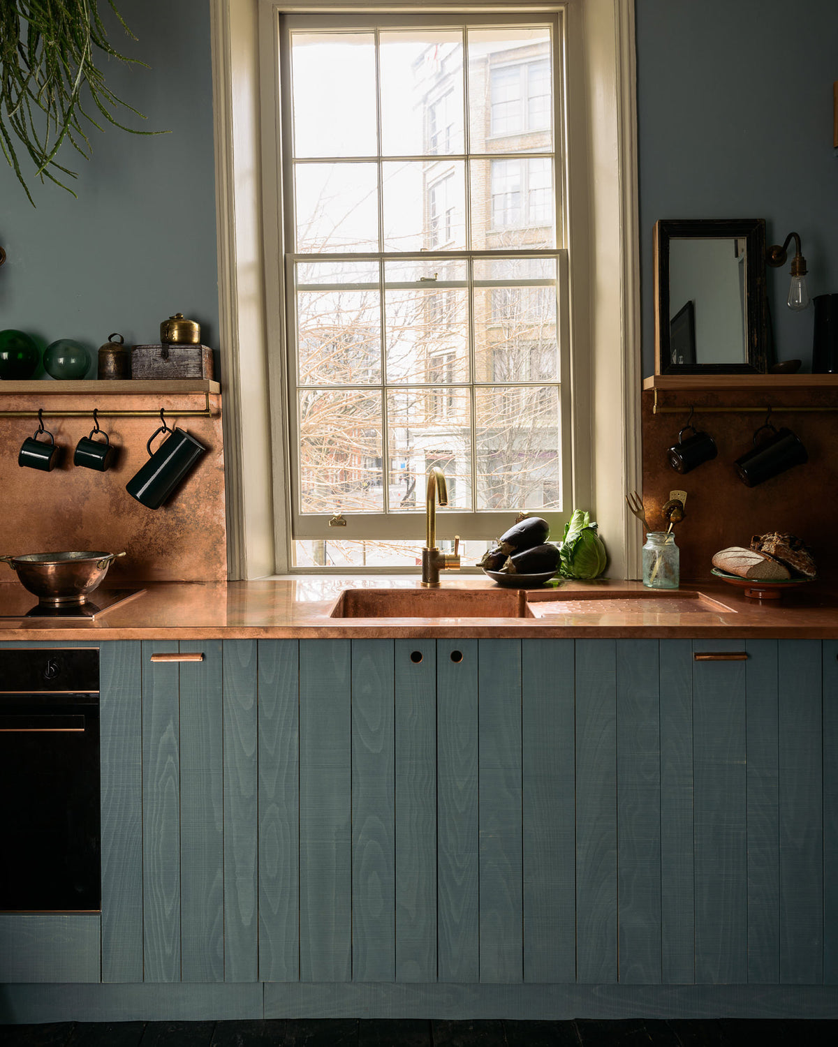 A sweet and simple Clerkenwell blue Sebastian Cox kitchen features a short cooker and sink run with a copper worktop and splashback, a single copper sink, an electric cooker, and shelves with brass hanging rails. Adorned with artisan ceramics, vintage bric-a-brac, aubergines, a sweetheart lettuce, loaves of bread, hanging plants, a framed quirky illustration, and bare wall lights, this kitchen creates a glamorous and practical space.