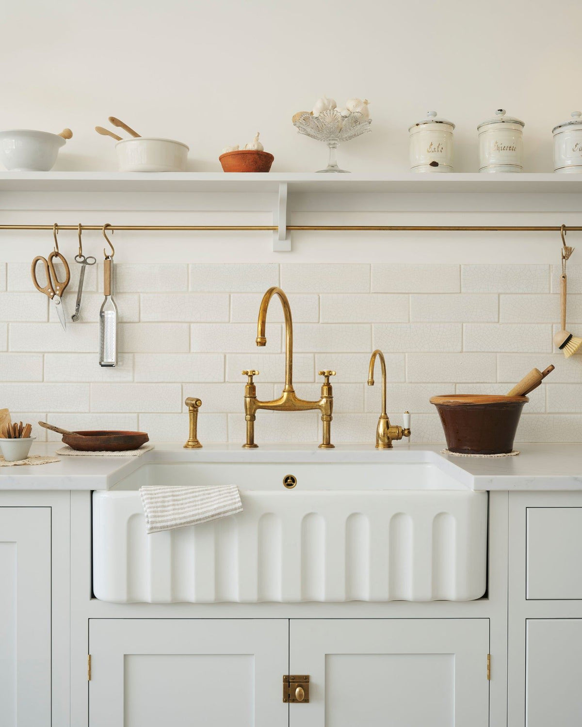 A close-up view of a fluted Belfast sink with white painted Shaker-style cupboard and an aged brass mixer tap. A simple shelf with a brass hanging rail and a splashback of white metro wall tiles with delicate crazing sits behind the tap. TEST