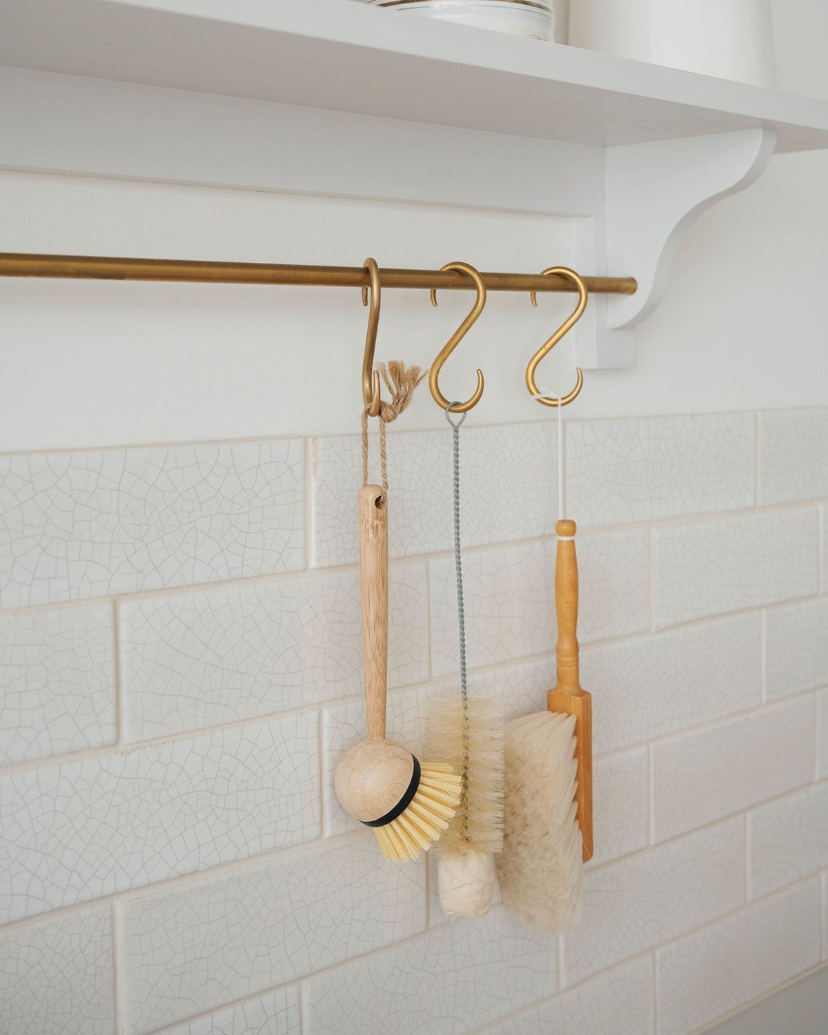 An oblique close-up view of an assortment of kitchen utensils hanging from a brass rail in front of white metro wall tiles with delicate crazing. TEST