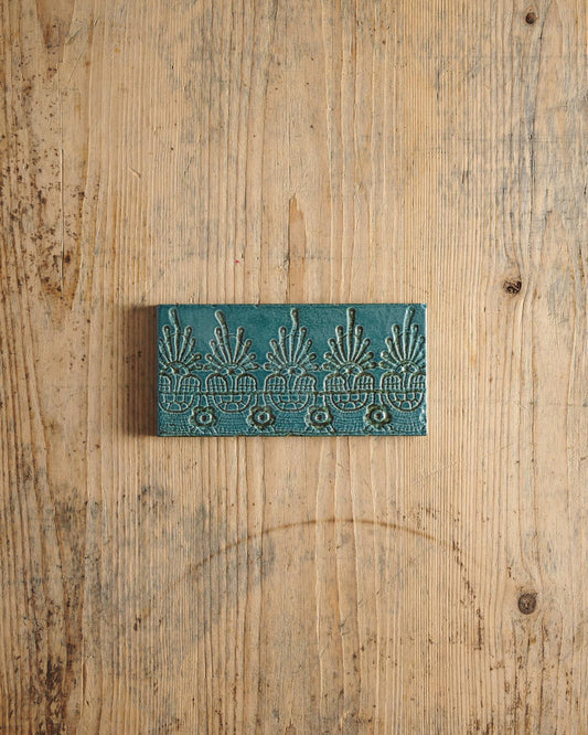 A glossy teal metro tile with a decorative lace pattern on a rustic wooden surface. | Flanders Metro