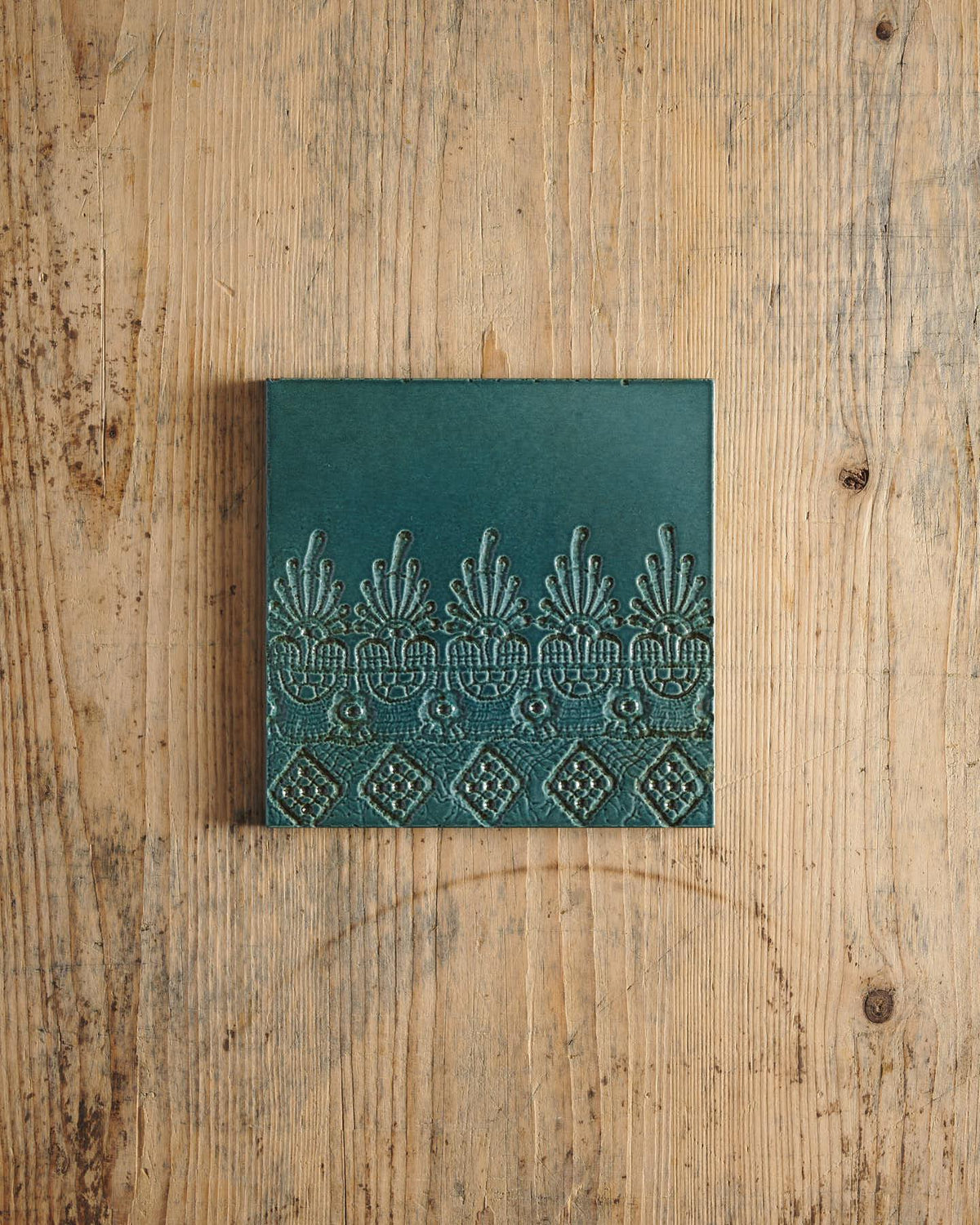A glossy teal square tile with a decorative lace pattern on a rustic wooden surface. | Flanders Square TEST