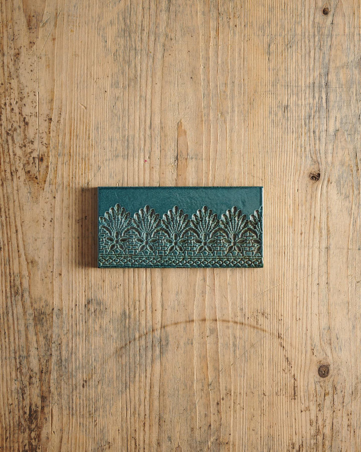 A glossy teal metro tile with a decorative lace pattern on a rustic wooden surface. | Flemish Metro TEST