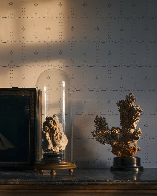 Warm light from a sunset is falling across a delicate patterned wallpaper with the shadow of a window frame visible behind antique ornaments on a table.|Petite Ditsy Delft in Blue